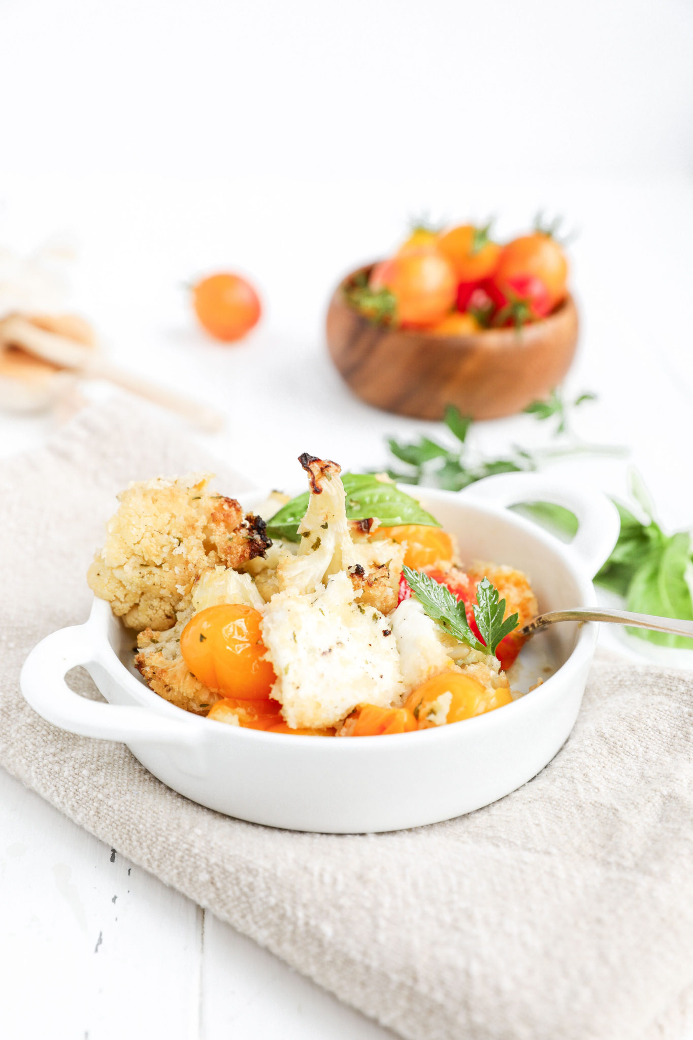 An individual bowl of cauliflower caprese casserole on a tan napkin with a wooden bowl of garden tomatoes behind it and fresh herbs scattered. 