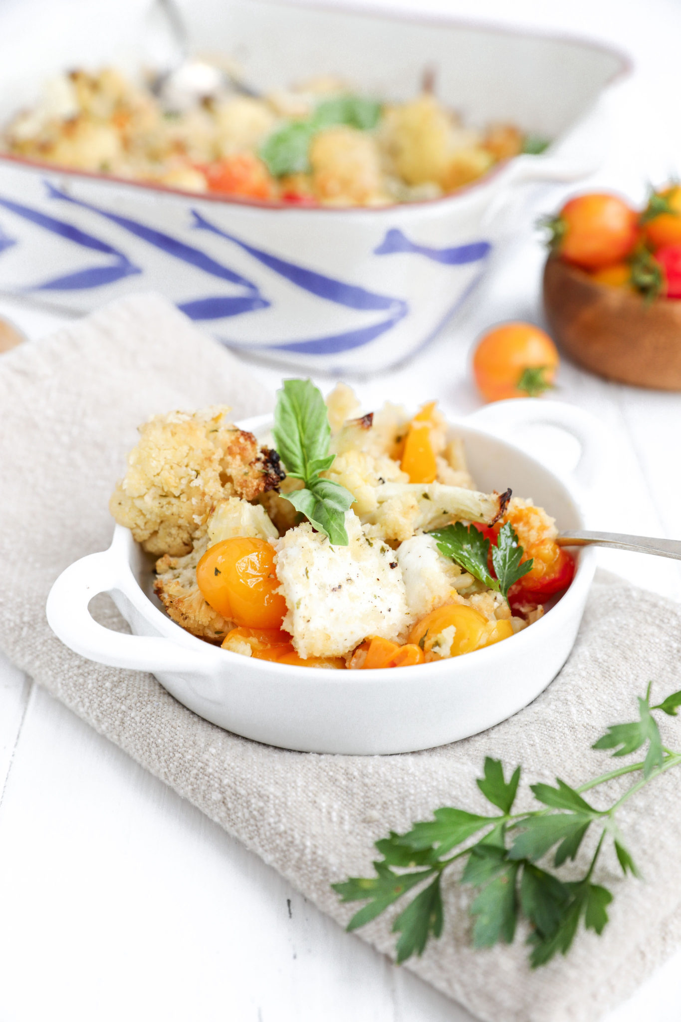 A small white bowl of cauliflower caprese casserole with a large dish of the casserole behind it. 