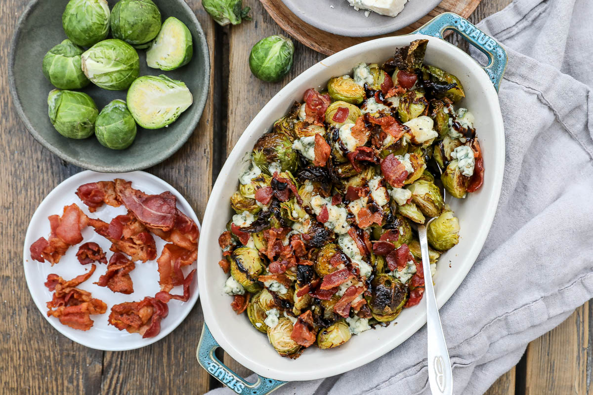 An overhead image of Brussels sprouts with bacon, blue cheese and balsamic drizzle in a casserole dish with a side plate of bacon and brussels sprouts.