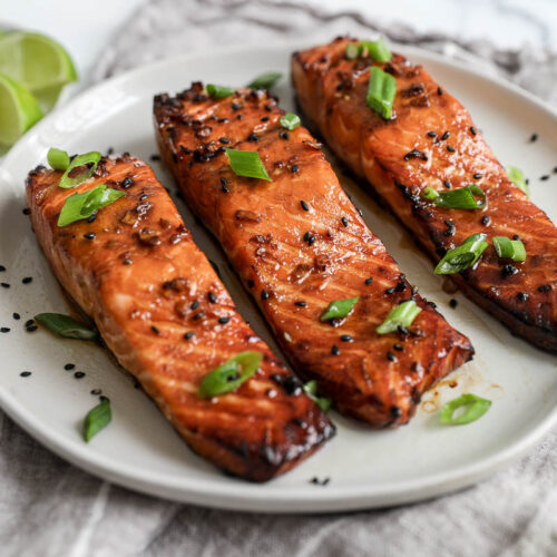 An image of 3 cooked salmon filets with scallions on a white plate.