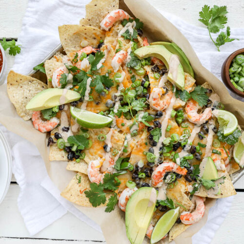 An image of shrimp nachos on a sheet pan on a white table with salsa and scallions.