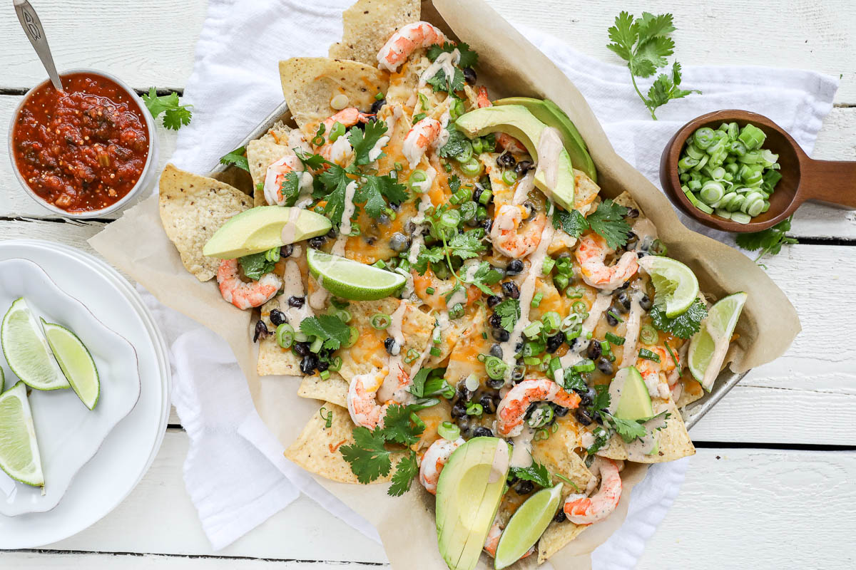 An image of shrimp nachos on a sheet pan on a white table with salsa and scallions.