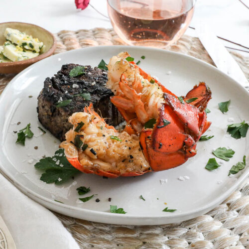 An image of lobster tails and filet mignon on a white plate with a glass of wine.