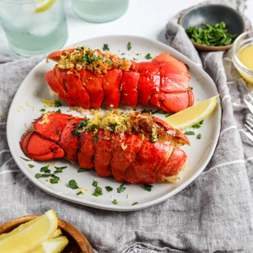 An image of two lobster tails on a white plate stuffed with panko crumbs on a grey and white napkin.