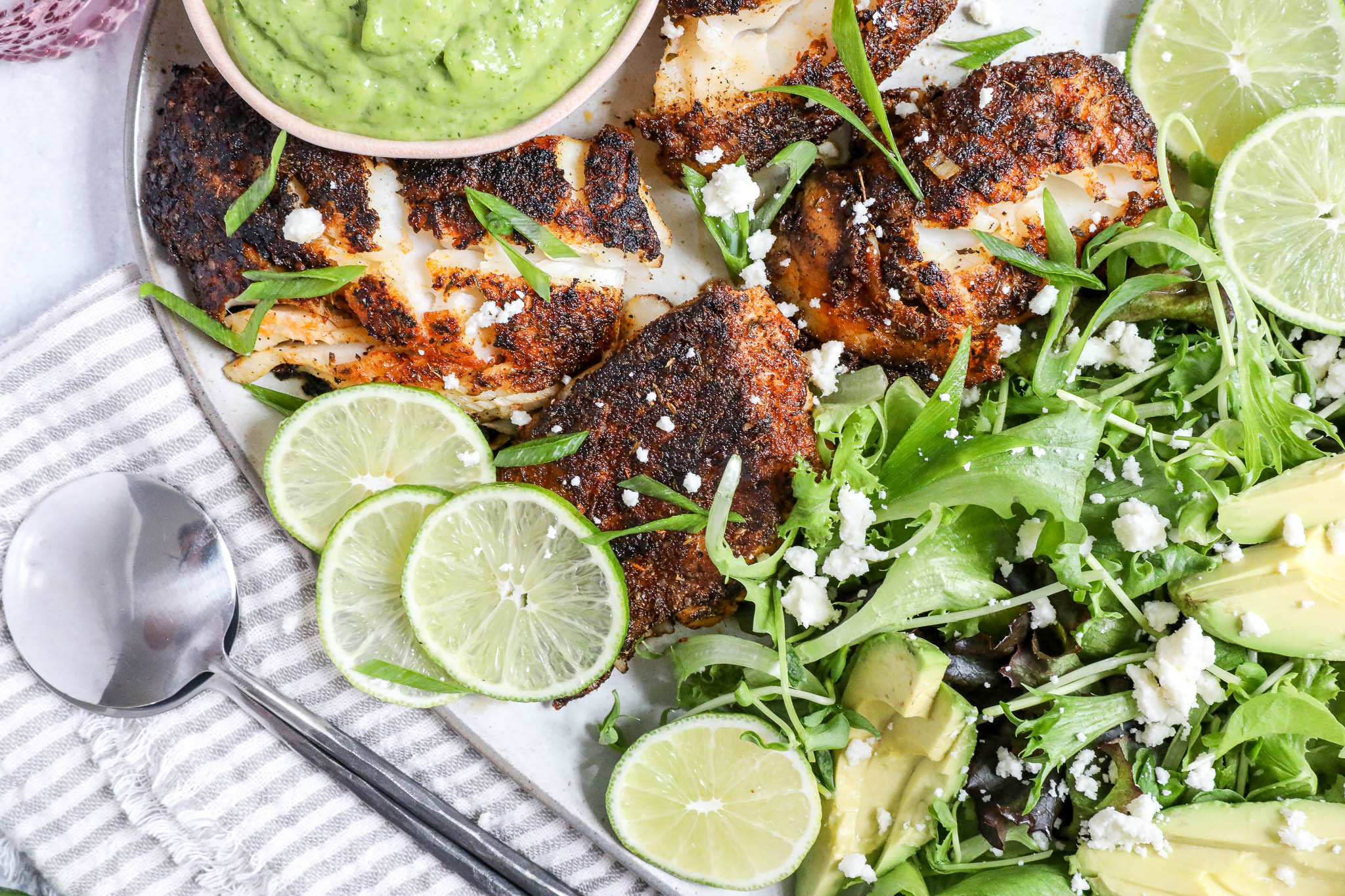 An overhead image of blackeend cod with a spring salad and lime wedges on a white platter with a grey striped napkin.
