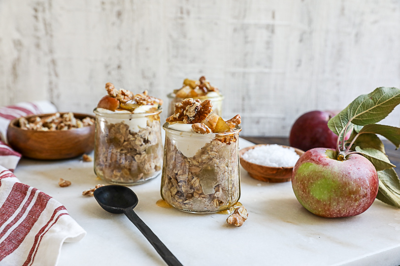 An image of jars of apple pie overnight oats topped with yogurt, apples, walnuts and honey served with a black spoon on a white slab of marble. 