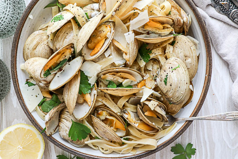 An overhead image of a large tan bowl of clams with pasta and parsley with a lemon on the side. 