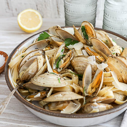 An image of clam pasta in a bowl with glasses of water and a lemon in the background.