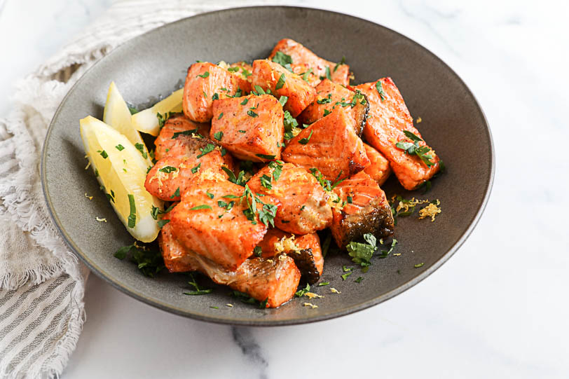 An image of air fried salmon bites with lemon zest and parsley in a black bowl with lemon wedges.