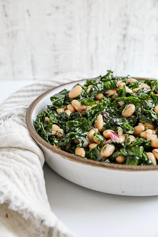 A close up image of a kale and white bean salad in a white bowl with a grey striped napkin.
