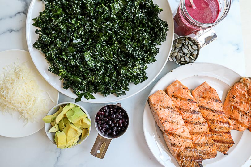 Ingredients to make a salmon kale salad laid out on a white counter, including: kale, avocado, wild blueberries, salmon, pumpkin seeds and wild blueberry vinaigrette.