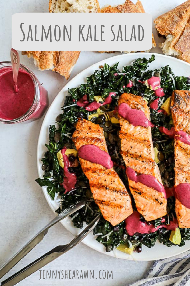 An image of a kale salad topped with grilled salmon and drizzled with a wild blueberry vinaigrette served on a white plate with more vinaigrette on the side and a torn baguette. 