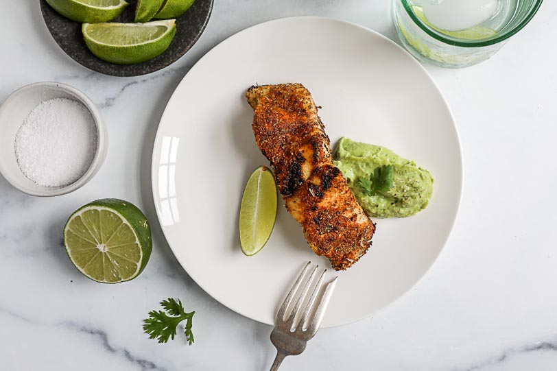 An overhead image of a filet of blackened grouper on a white plate with avocado crema and a lime wedge.