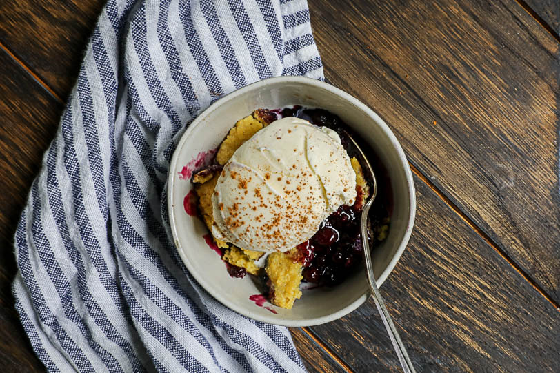 An image of a white bowl with wild blueberry cobbler and ice cream on top on a wooden table with a striped napkin.
