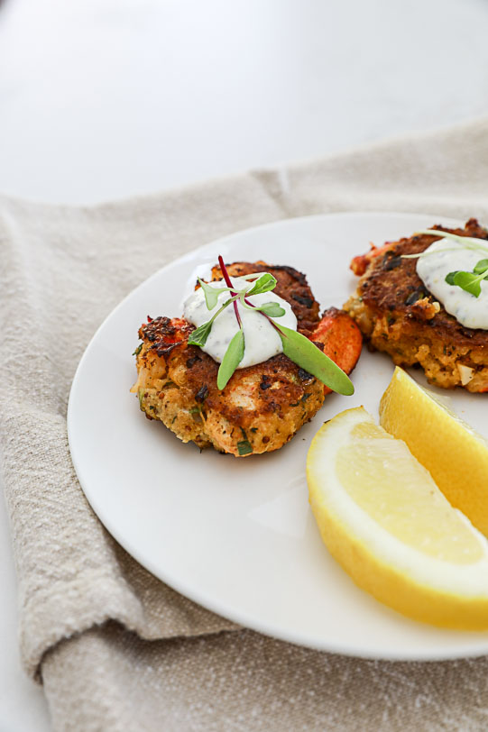 An image of 2 lobster cakes on a white plate with lemon wedges. 