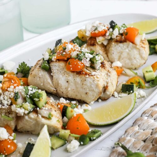 An image of grilled striped bass on a white platter topped with salsa and served with lime wedges.