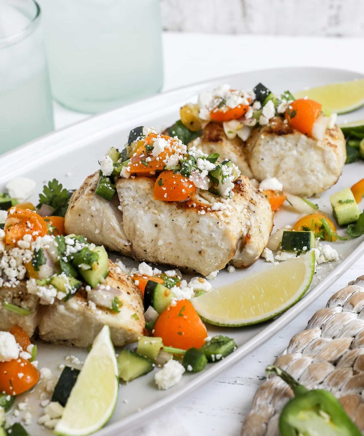 An image of grilled striped bass on a white platter topped with salsa and served with lime wedges. 