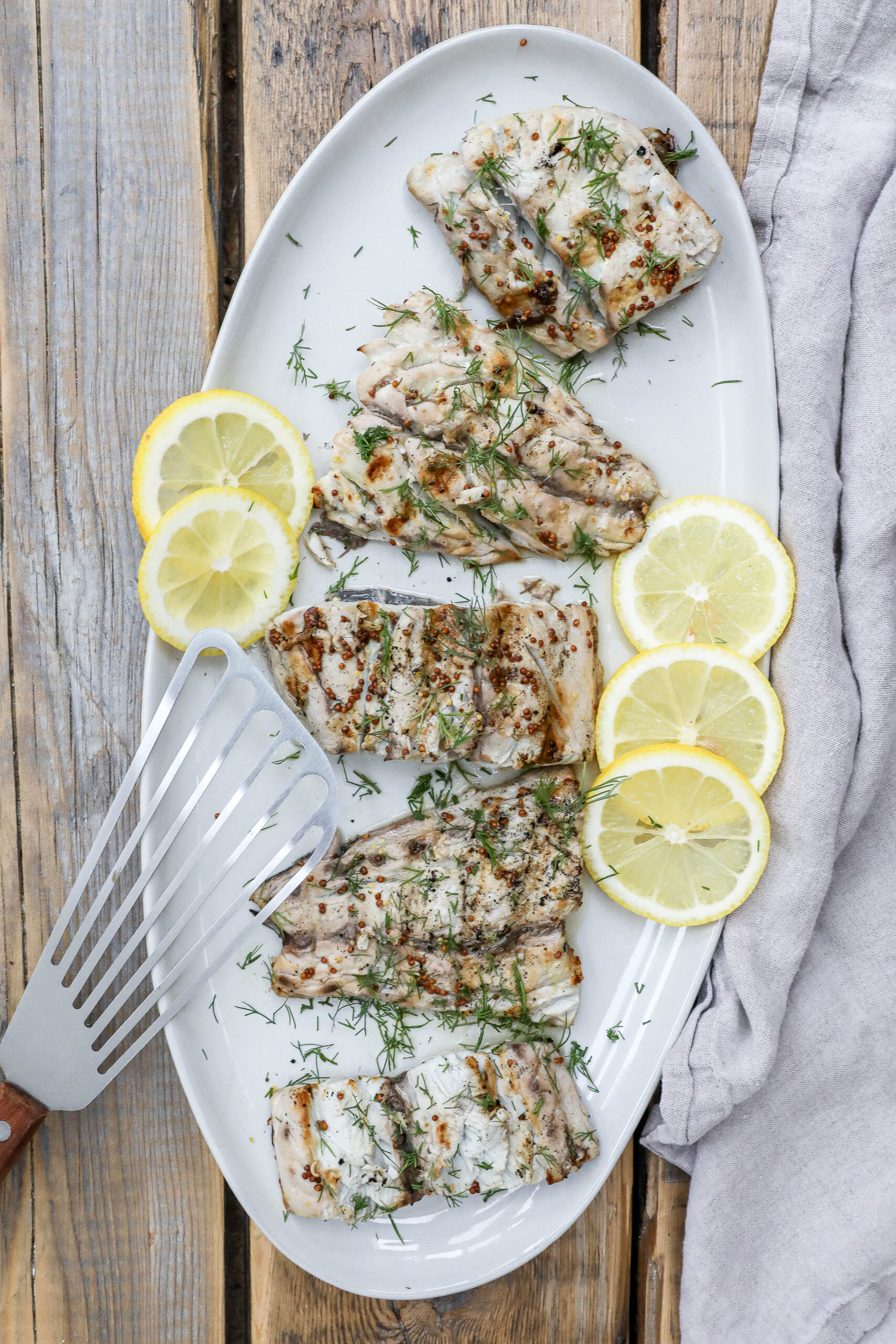 An overhead image of grilled bluefish fillets with lemon slices on a white plate with a fish spatula. 