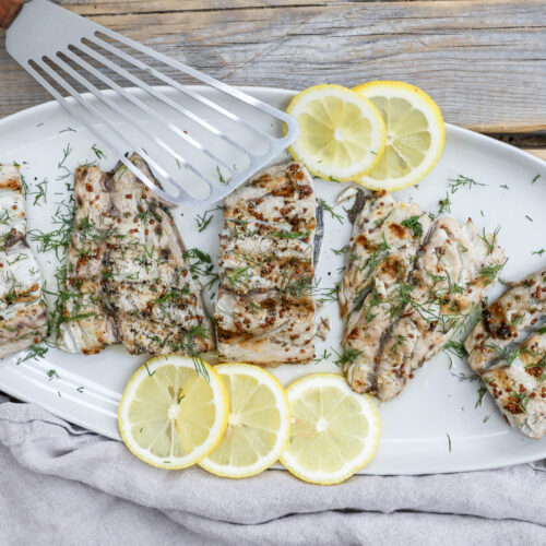 An image of grilled bluefish with lemon and dill on a white plate with a fish spatula.