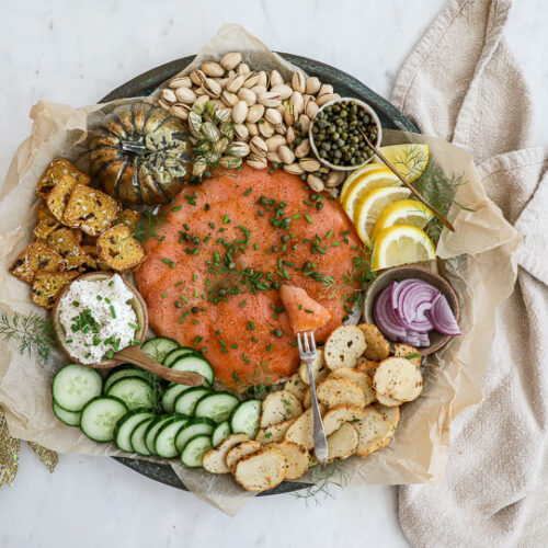 An overhead image of a smoked salmon board with cucumbers, cream cheese, crackers, red onion, lemon, pistachios and more.