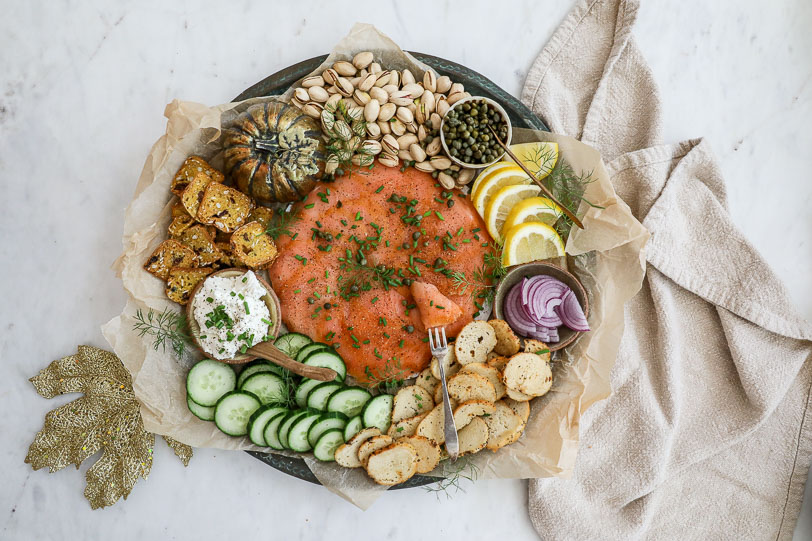 An overhead image of a smoked salmon board with cucumbers, cream cheese, crackers, red onion, lemon, pistachios and more.