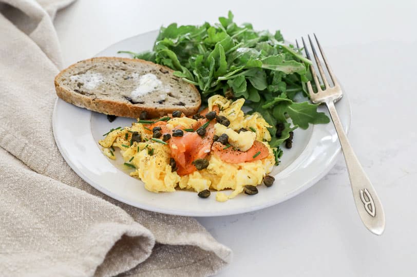 An image of smoked salmon with eggs on a white plate with toast and arugula. 