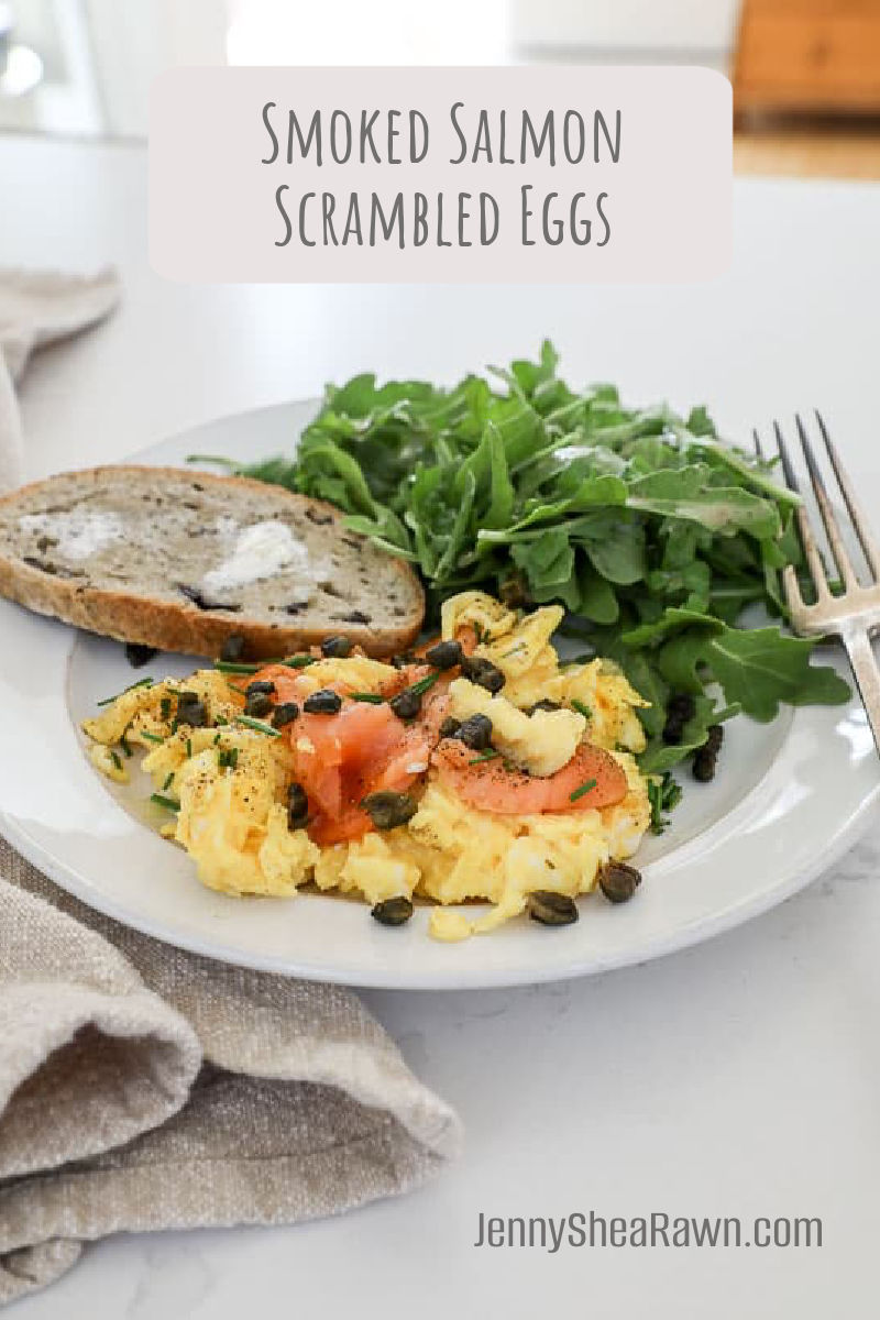 An image of smoked salmon scrambled eggs on a white plate with toast and an arugula salad.