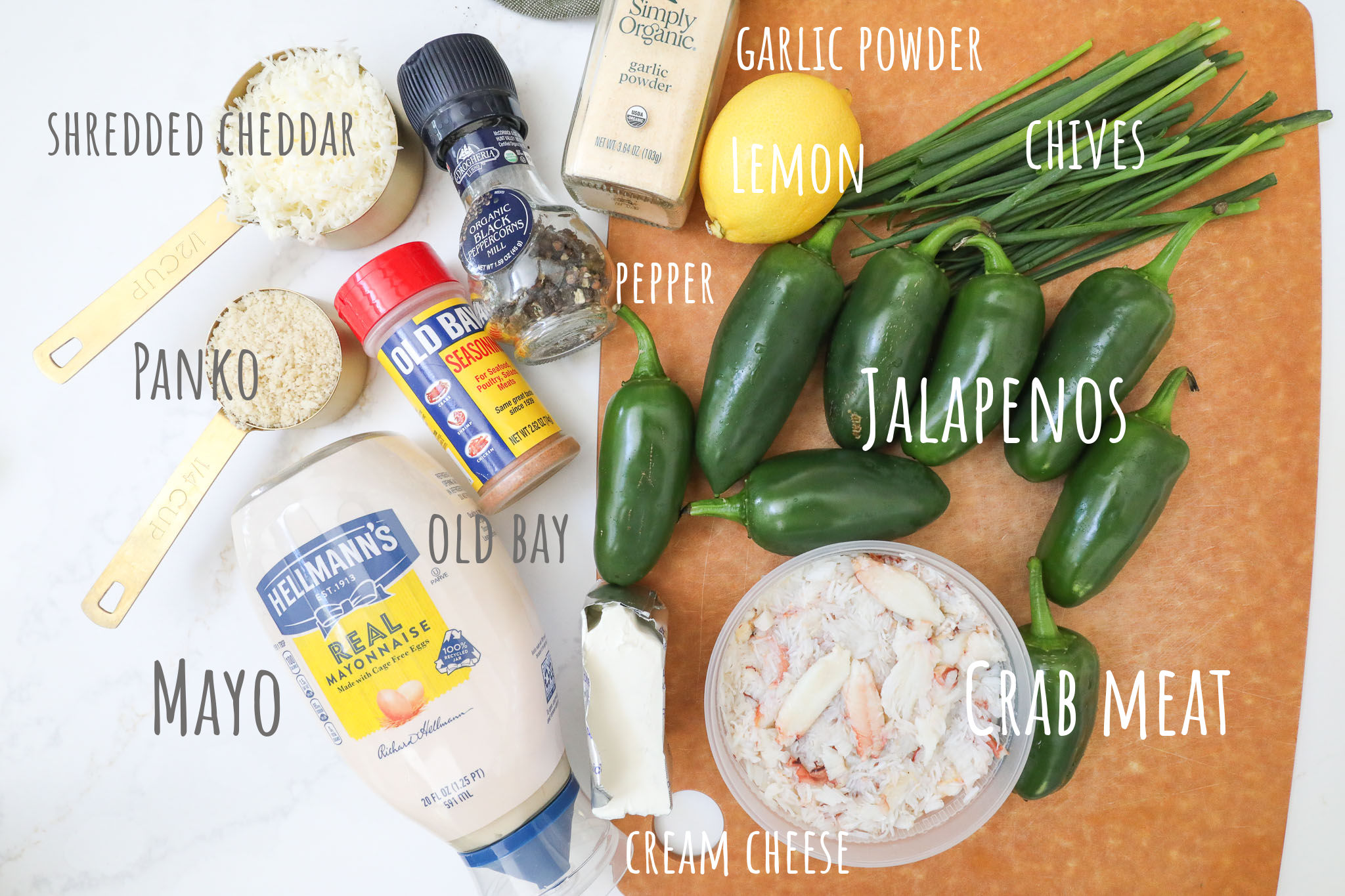 Ingredients to make crab stuffed jalapenos laid out on a cutting board on a white counter. Crab, panko, peppers, chives, lemon, old bay, mayo, cheddar, cream cheese, garlic powder and pepper.