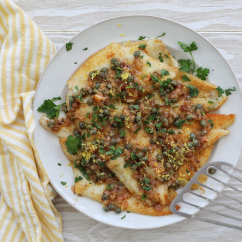 An image of sole piccata fillets on a large white plate with a fish spatula to the right and a white and yellow napkin to the left.