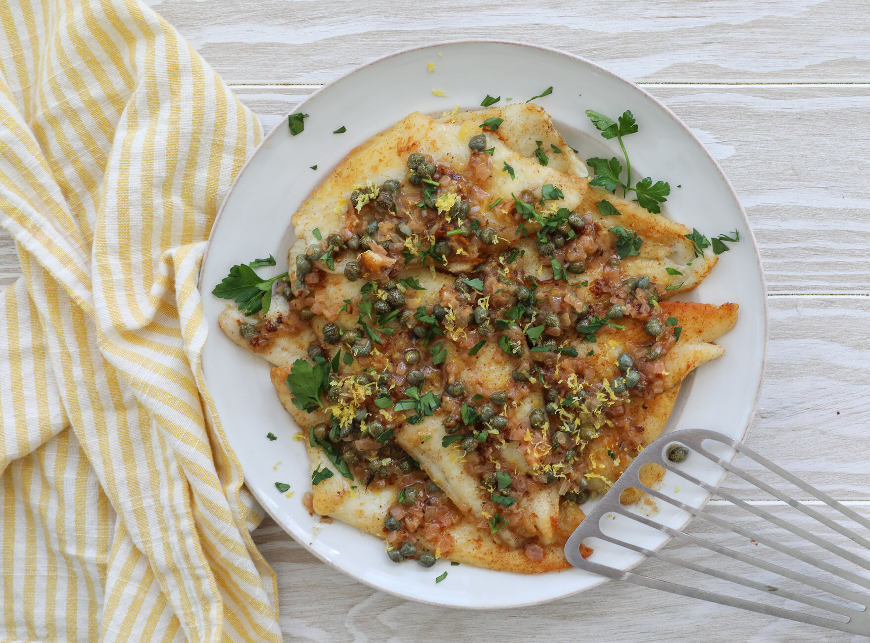 An image of sole piccata fillets on a large white plate with a fish spatula to the right and a white and yellow napkin to the left.