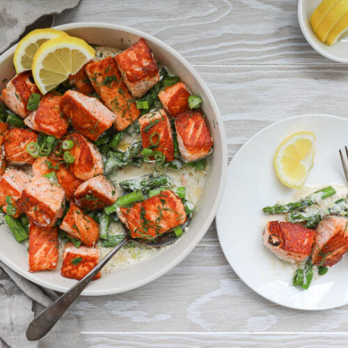 An overhead image of salmon bites in cream sauce with asparagus and a small plate on the side.