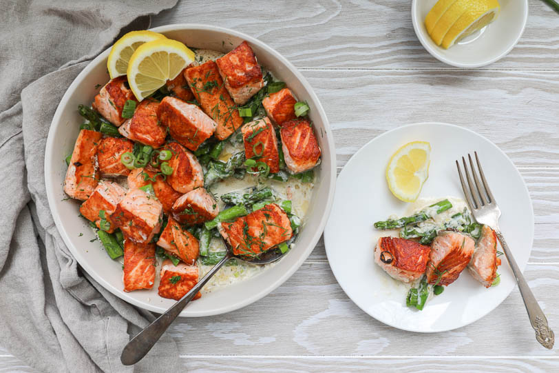 An overhead image of salmon bites in cream sauce with asparagus and a small plate on the side.