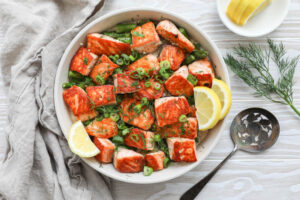 An overhead image of salmon bites in a white bowl with a cream sauce, asaparagus and lemon slices.